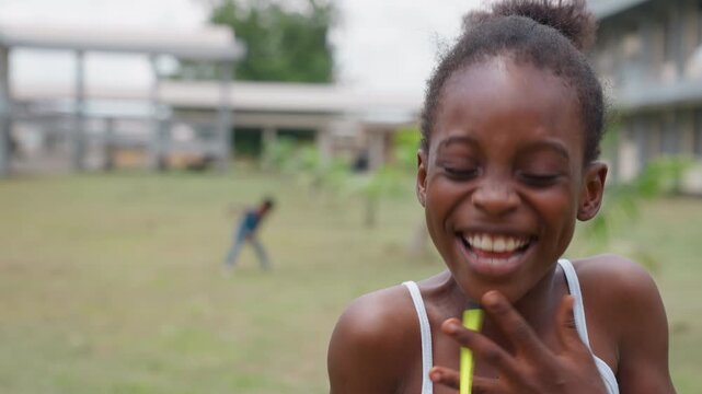 Girl smiles while balancing yellow pencil on face. Outdoors near building and grassy area. Child hair in top knot smile outdoor school grass white shirt