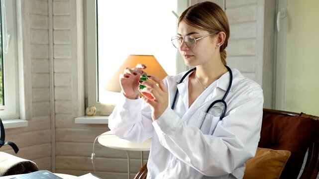 Young female doctor inspects green medicine bottle to avoid medication error risk while seated in room, carefully checking liquid pharmaceutical dosage before writing patient prescription.