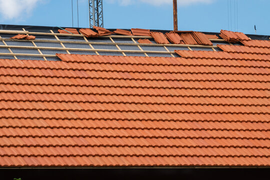 a house roof undergoing tiling renovation with red clay tiles partially installed over wooden battens and protective black membrane under a dramatic cloudy sky.
