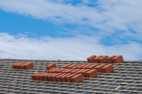 Bundles of red clay roof tiles stacked across wooden battens and protective underlayment during a roof installation against a blue, cloudy sky.