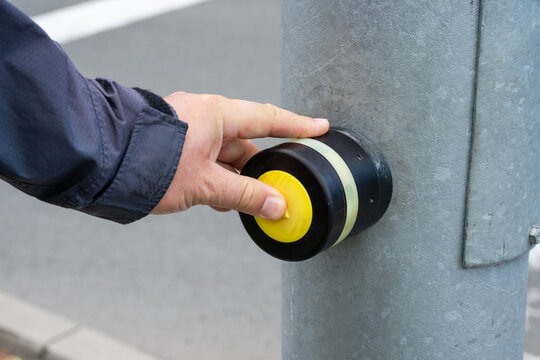 a person's hand pressing a round yellow pedestrian crossing button mounted on a metal street pole to request a walk signal.