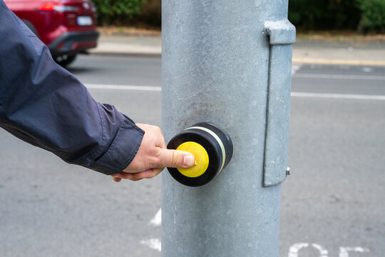 a person's hand pressing a round yellow pedestrian crossing button mounted on a metal street pole to request a walk signal.