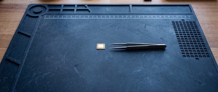 Tiny microchip component held by tweezers on black anti static mat, close up of electronic part for repair and manufacturing, precision assembly workspace.