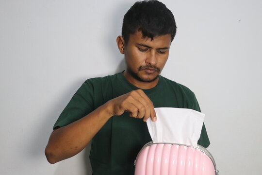 Young man pulling white paper tissue from pink container against white wall