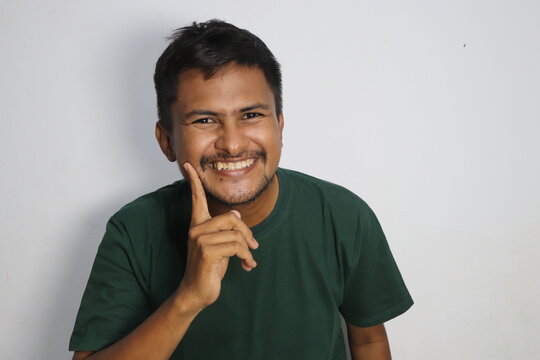Young man smiling and pointing finger on cheek against white background