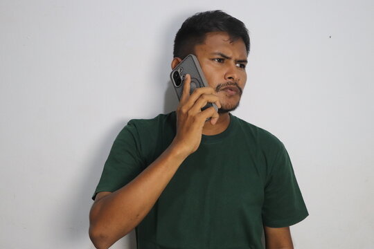 Young man talking on smartphone wearing green t shirt against white wall