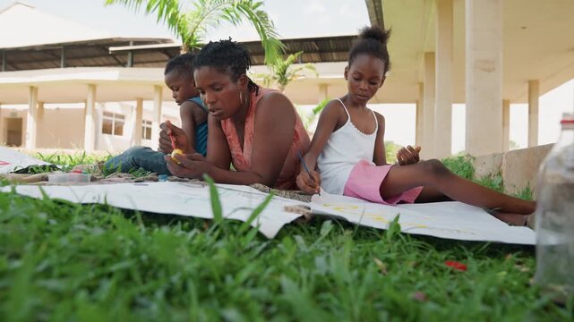 Family members lie together outdoors creating art with paint. woman assists young girl in drawing on large sheets while another child plays nearby. scene is set under open structure