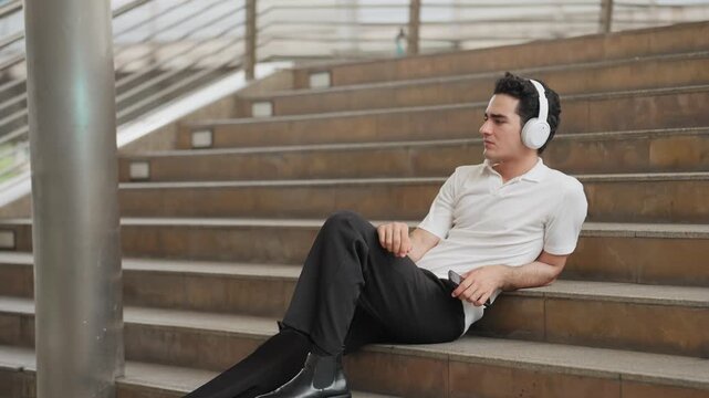Man sits relaxed on urban stairs. He wears white headphones, eyes closed. Soft ambient light bathes the stone steps. He appears calm