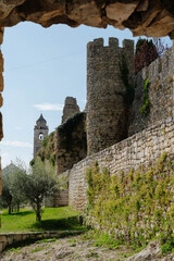 Stone castle walls and a crenellated tower in Montemor o Velho Portugal rise beyond olive trees and a church bell tower framed by an archway. Medieval fortification for travel and heritage tourism. © Renata
