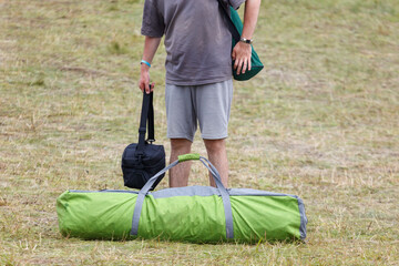 Man stands beside rolled green camping bag. Grassy field surrounds the scene. Man holds black bag and green shoulder bag. © schankz