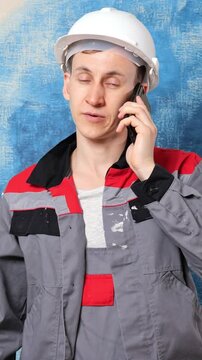 Young male contractor in a protective helmet and stained work uniform seriously talking on his smartphone, managing construction site issues and coordinating tasks with his team