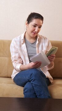 Excited brunette woman opening a white envelope filled with cash, joyfully counting the dollar bills and celebrating her financial gain while relaxing on a comfortable couch at home