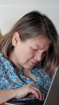 Woman types on laptop at home, focused on screen while seated by sofa. Home computing task continues as woman works with steady concentration, using keyboard and trackpad during quiet indoor moment.