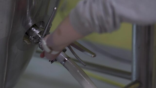 Brewery worker adjusts stainless steel tank valve and braided hose during a brewing process in an industrial beer production facility