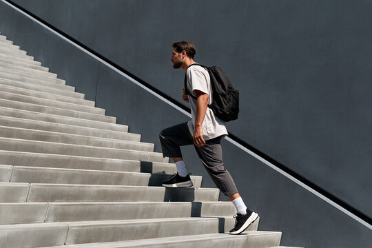 Young man ascends concrete stairs with backpack. Sunlight casts sharp shadows across steps and wall. He moves forward, focused, unbothered by ascent