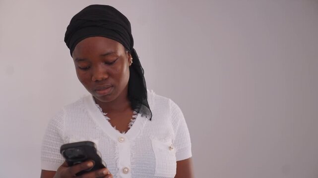 Woman in white frayed collared shirt with pearl button details and black headwrap holding her smartphone. Woman maintains focus on device against plain background