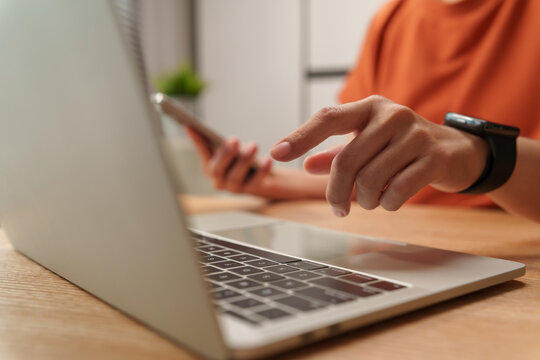 Woman using a smartphone beside a laptop while reaching toward the computer, capturing digital multitasking, connectivity and modern mobile productivity.