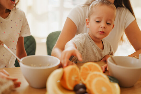 Little toddler boy reaching for fresh orange slice on plate during family breakfast in kitchen with mother and sister sitting at wooden table at home
