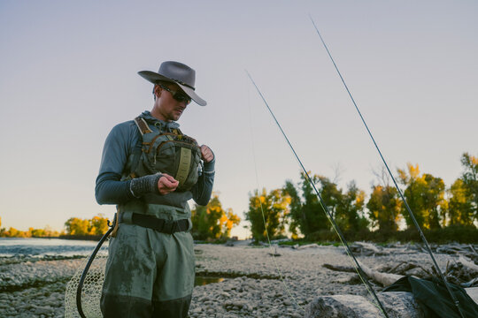 Fly fisherman organizes fishing gear beside a rocky riverbank.