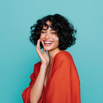 Curly haired woman smiling with moisturizer on face wearing red dress on teal background for beauty campaign and skincare wallpaper design