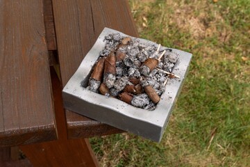 Fototapeta premium High angle view of a square stone ashtray filled with smoked cigarette butts and ash placed on an outdoor wooden table in daylight