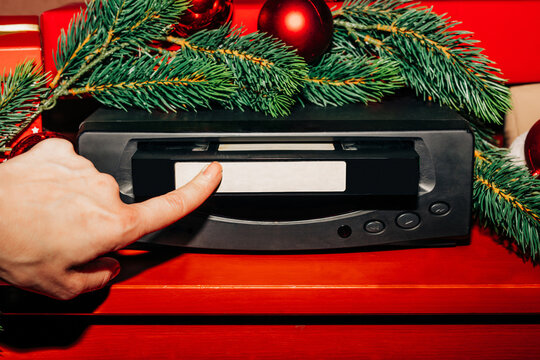 female hand inserts a Christmas-themed VHS tape into a VCR decorated with fir branches and a red bauble, ready to watch nostalgic home videos from the 90s