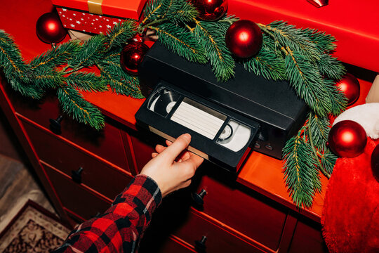 female hand inserts a Christmas-themed VHS tape into a VCR decorated with fir branches and a red bauble, ready to watch nostalgic home videos from the 90s