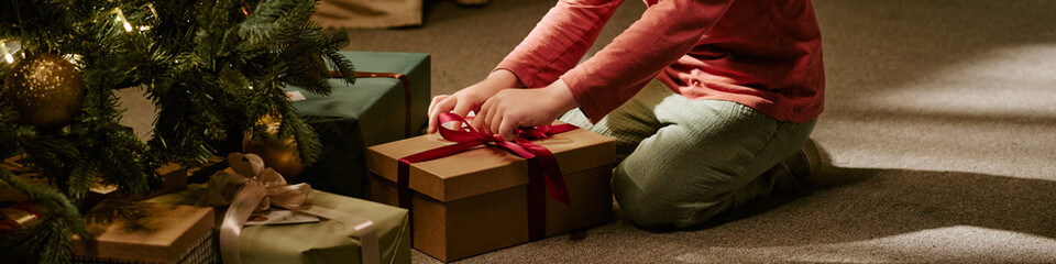 Fototapeta premium Header of Caucasian child opening gift box near Christmas tree at home. Image suitable for holiday marketing campaigns, seasonal retail promotions, family lifestyle advertising