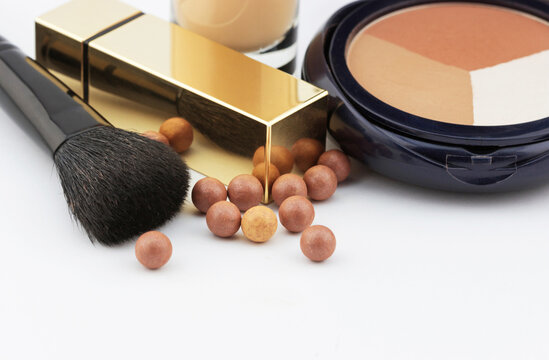 Close view of luxury cosmetic products including a makeup brush, gold lipstick case, bronzing pearls, liquid foundation, and face powder compact on white