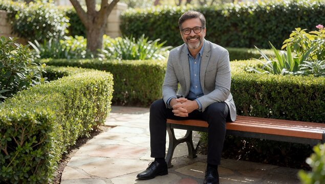 Happy middle aged man wearing glasses and gray blazer sitting on a wooden bench in a lush green garden during a bright sunny day
