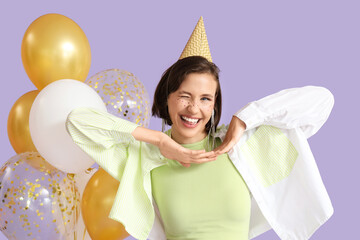 Fototapeta premium Birthday celebration and party joy, happy young woman wearing a festive hat winking with gold balloons on a purple background
