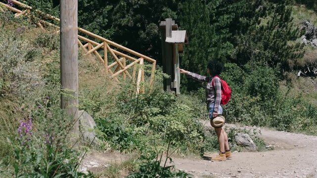 Hiker woman is walking down a dirt road approaching the trail map info wearing backpack on her back. She is wearing shorts and a hat