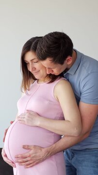 Loving Man Hugs Pregnant Woman From Behind While Holding Hands On Baby Bump Indoors. Caring Couple Looks Down With Affection, Enjoying Peaceful Maternity Moment Beside Bright Wall In Studio.