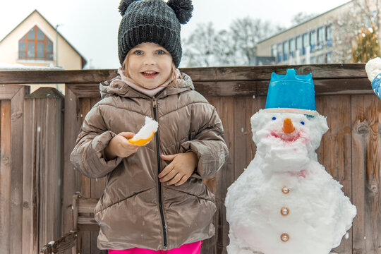 Happy child smiling at camera holding snack next to funny snowman on wooden balcony outdoor winter activity portrait joy authentic emotion child care seasonal promotion