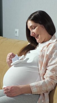 Happy Future Mother Rests On Home Sofa While Playing With Small Newborn Socks On Belly. Delighted Woman Holds Soft White Booties Against Round Tummy, Experiencing Pure Maternal Joy Inside.