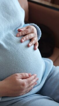 Smiling Expectant Mother Holding Pregnant Belly With Both Hands Indoor. Joyful Brunette Woman Sitting Comfortably Indoors Enjoys Serene Quiet Moment Of Bonding With Baby Before Childbirth.