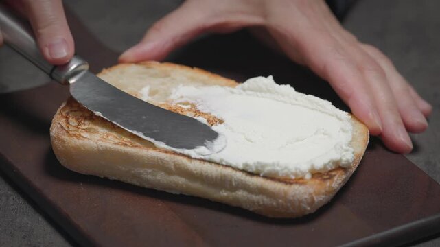 chef spreads fresh Italian ricotta cheese on slice of toasted ciabatta bread on wooden kitchen board set on black tabletop. The process of preparing Italian tomato and ricotta bread; food closeup