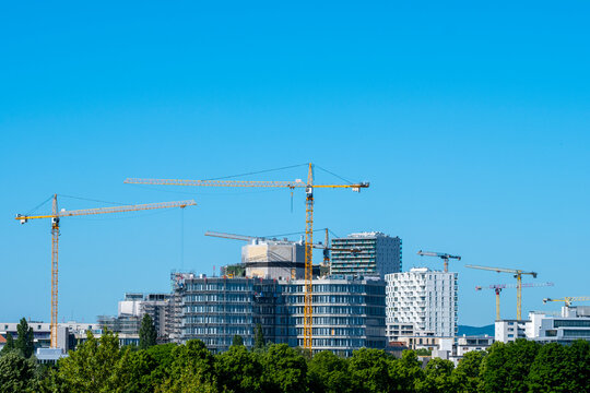 Construction cranes rise above modern buildings and trees in Vienna Austria with an urban skyline under blue sky during clear daytime conditions