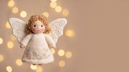 Naklejka premium Handmade angel ornament with curly hair, knitted cream dress, embroidered flowers, and white wings suspended against warm golden bokeh lights