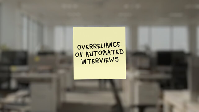 Yellow sticky note with the inscription "OVERRELIANCE ON AUTOMATED INTERVIEWS" in the foreground, superimposed on a blurred background showing an open-plan office workspace.