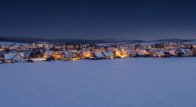 Winter twilight landscape over snowy field with illuminated village houses in a rural sunset scene