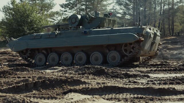 Side view of an armored tracked infantry fighting vehicle moving across muddy terrain surrounded by pine trees.