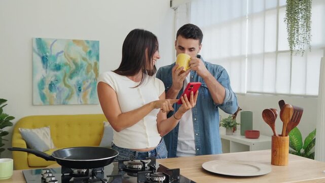 A cheerful duo engage with a red smartphone while preparing a meal in a sunny kitchen. They lean over a wooden counter with a pan, utensils, and a yellow mug nearby, exuding collaboration and