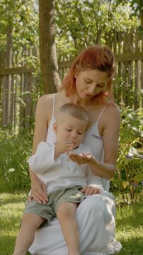 Vertical Screen: A woman and her little boy sit in the yard. They pick and eat ripe raspberries together. They share smiles and laughter as they enjoy their time outside.