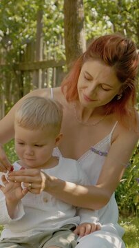Vertical Screen: A woman and her little boy sit in the yard. She places raspberries on his fingers while they laugh together. They are spending quality time on a sunny day.