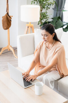 Senior digital lifestyle and remote work concept, mature Asian businesswoman with pearl necklace using laptop at home coffee table, representing active retirement, freelance, and online study.