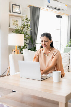 Senior digital lifestyle and remote work concept, mature Asian businesswoman with pearl necklace using laptop at home coffee table, representing active retirement, freelance, and online study.