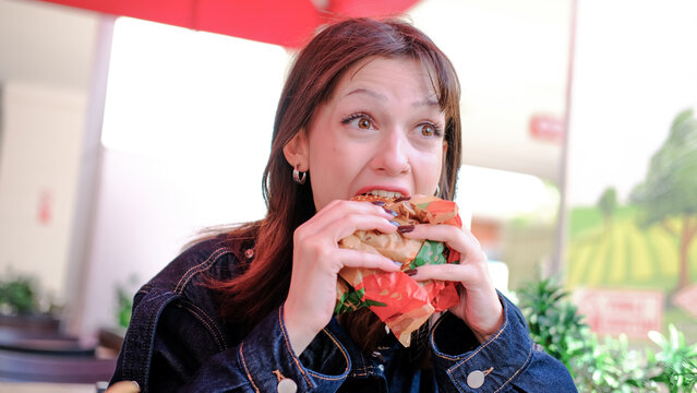 Young woman taking a big bite of a burger with lettuce and tomato at an outdoor cafe, holding it with both hands, long brown hair, dark denim jacket, red umbrella above, bright day, fast food