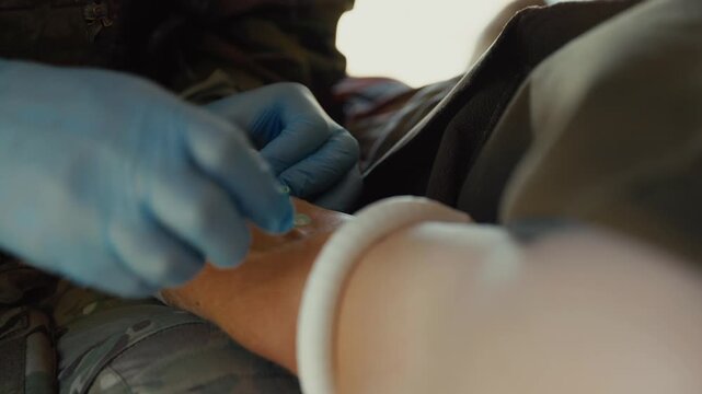 Close-up of a gloved medic inserting an IV catheter into a patient's arm at a tactical medical stabilization point.