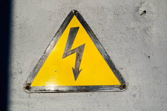 Industrial safety background with a weathered yellow triangular warning and a black lightning bolt symbol mounted on a grey metal surface, high voltage or electrical hazard warning concept
